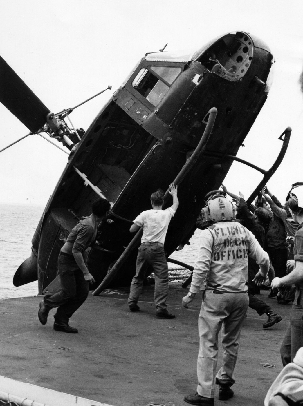 Czarno-biała fotografia: marynarze na pokładzie okrętu spychają śmigłowiec za burtę do morza; jeden ma na koszulce napis Flight Deck Officer.