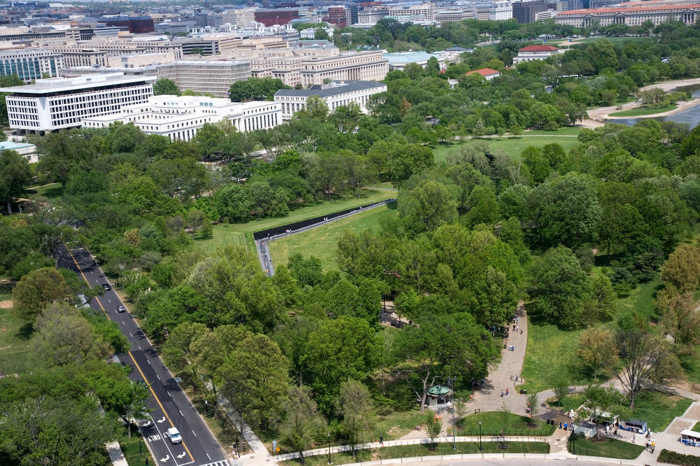 Widok z lotu ptaka na park z długą czarną ścianą w kształcie litery V wpuszczoną w trawnik; wzdłuż niej idą zwiedzający, wokół drzewa i gmachy rządowe.