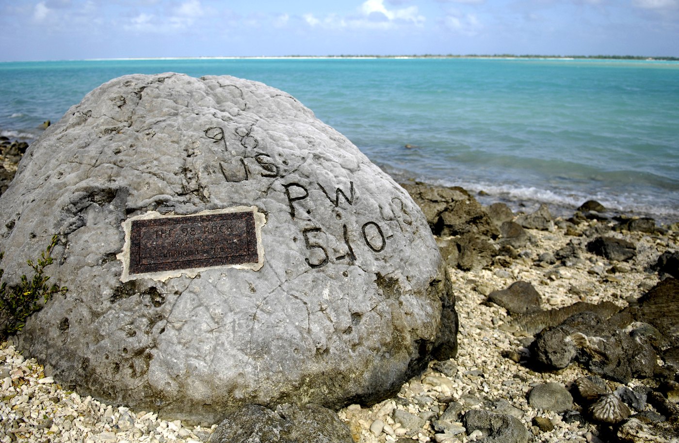 A large gray coral boulder on a shingle beach beside a turquoise lagoon, carved with the letters 98 US PW 5-10-43 and fitted with a small dark plaque