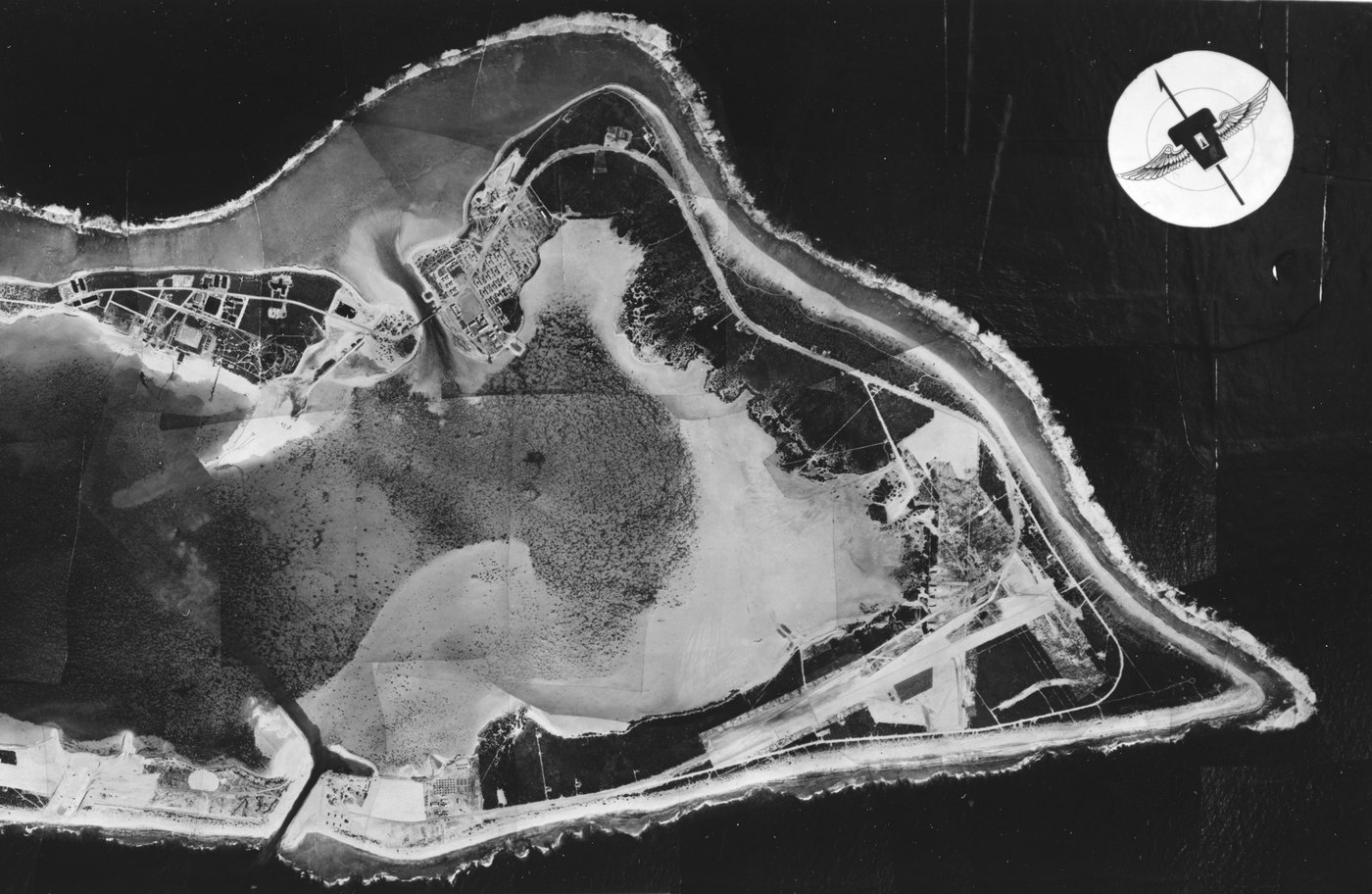 Vertical black and white photo mosaic of a V shaped atoll around a pale lagoon, with buildings on the islet at upper left, a triangular airstrip at lower right, white surf along the reef and a winged emblem with a north arrow at upper right