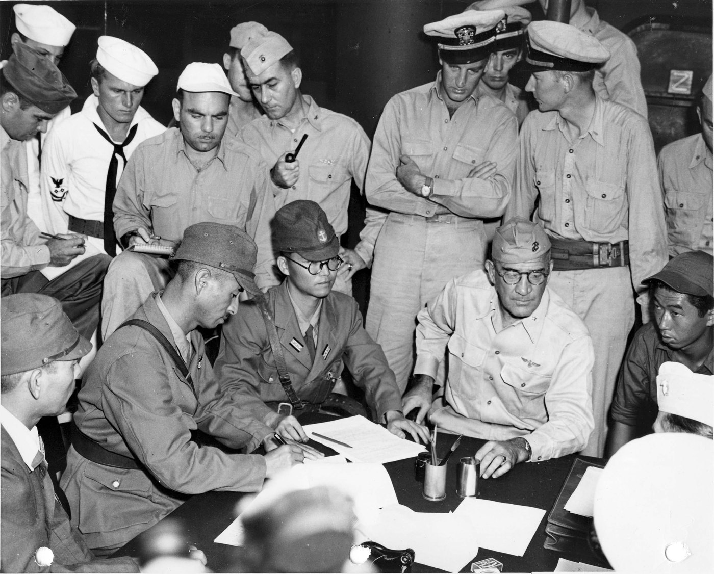 Japanese officers in field caps sit at a table signing papers in a crowded ship's compartment, facing a bespectacled American Marine general, with American officers and sailors in white caps standing behind them