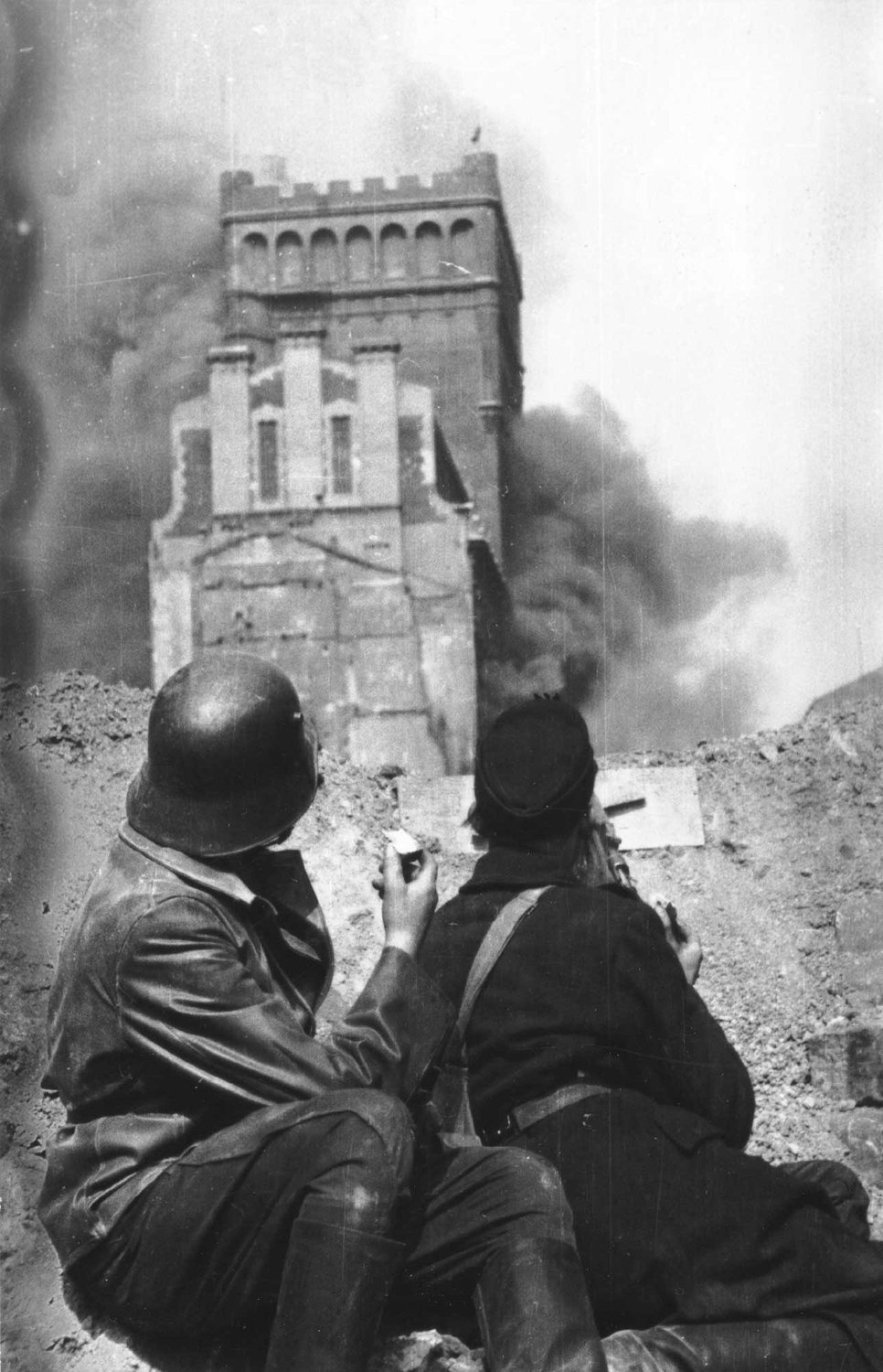 Two insurgents in helmets crouch on a barricade looking at a burning tower block