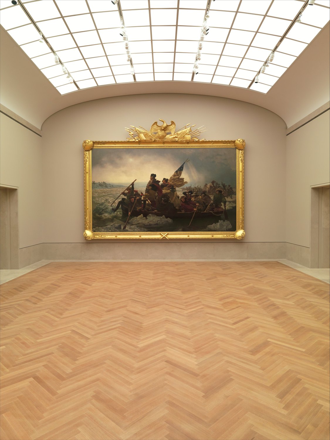 A large painting of Washington in a boat hanging alone on the end wall of a skylit museum gallery in a gilded frame topped by an eagle
