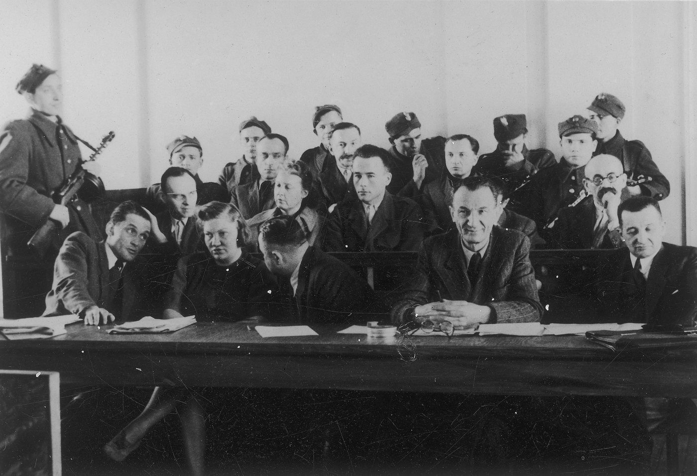 Black and white photograph of defendants seated behind tables in a courtroom, guarded by soldiers