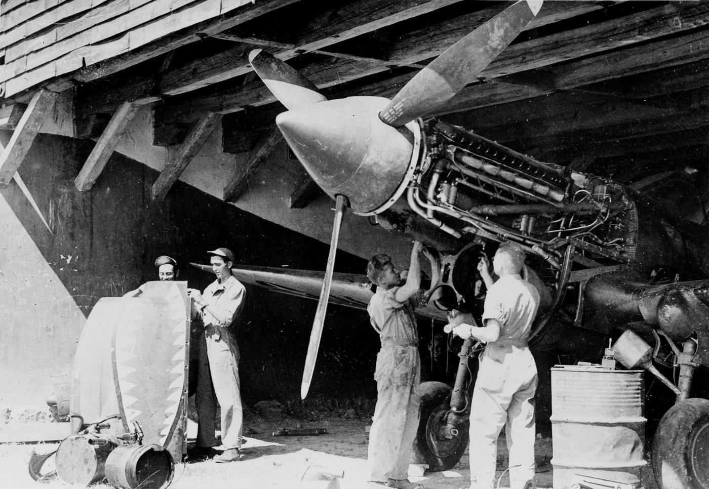 Mechanics working on the engine of a P-40 fighter under a wooden shelter