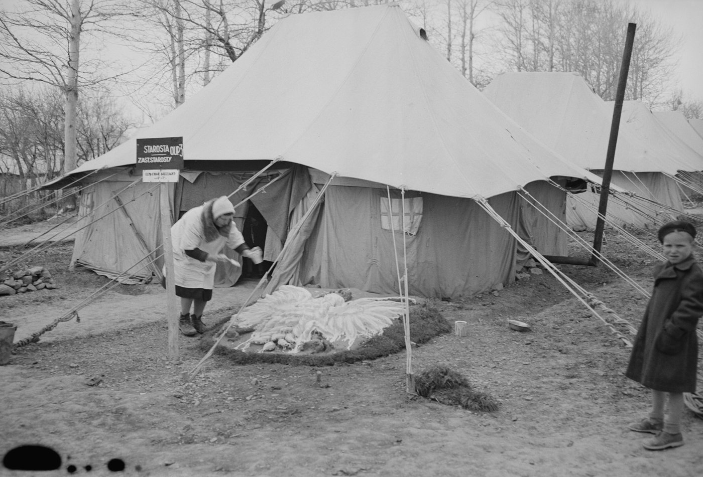 A woman bending over a design laid out on the ground in front of large tents in a refugee camp