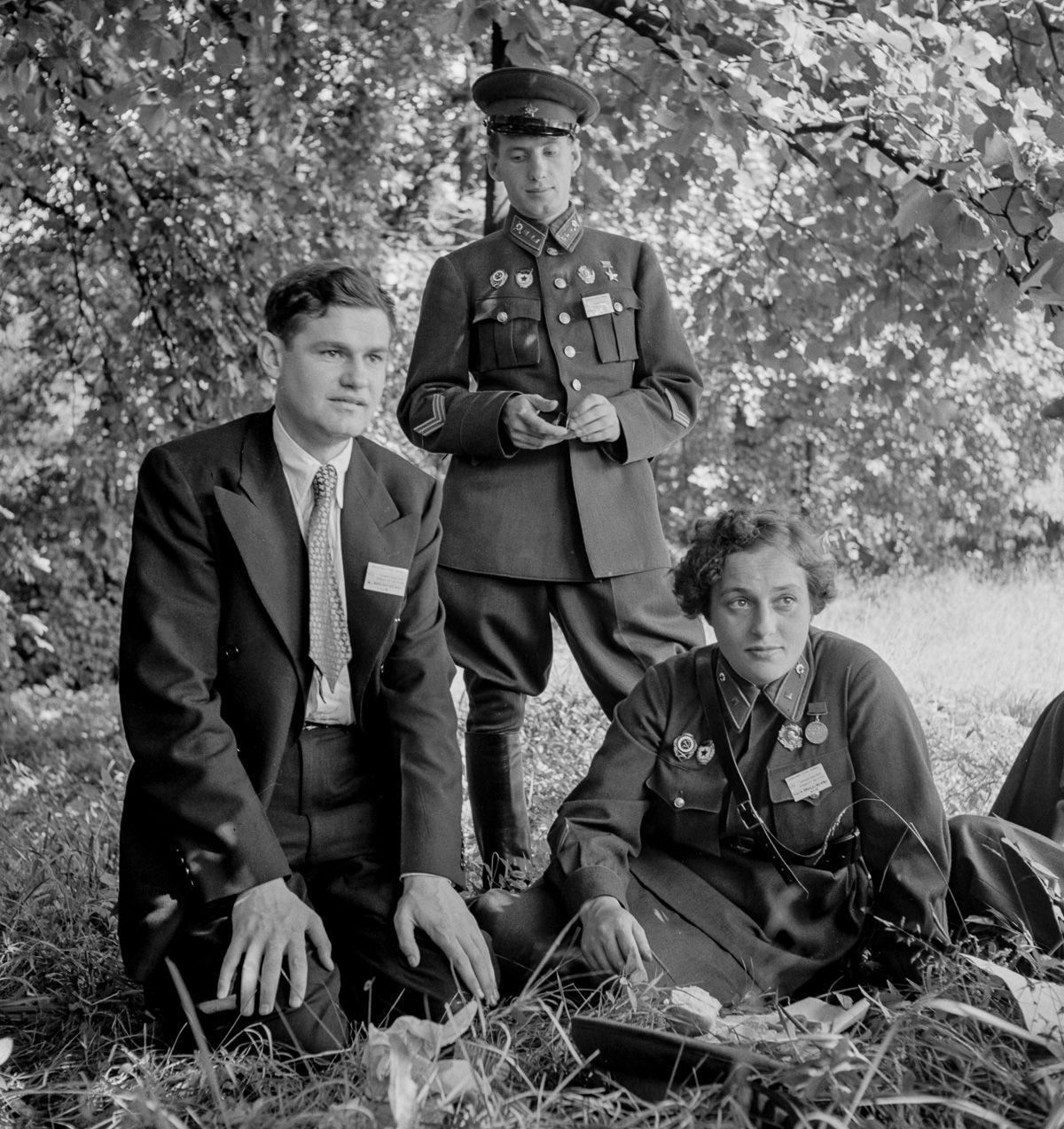 Lyudmila Pavlichenko in uniform sitting on the grass beside a man in a suit, with an officer standing behind them