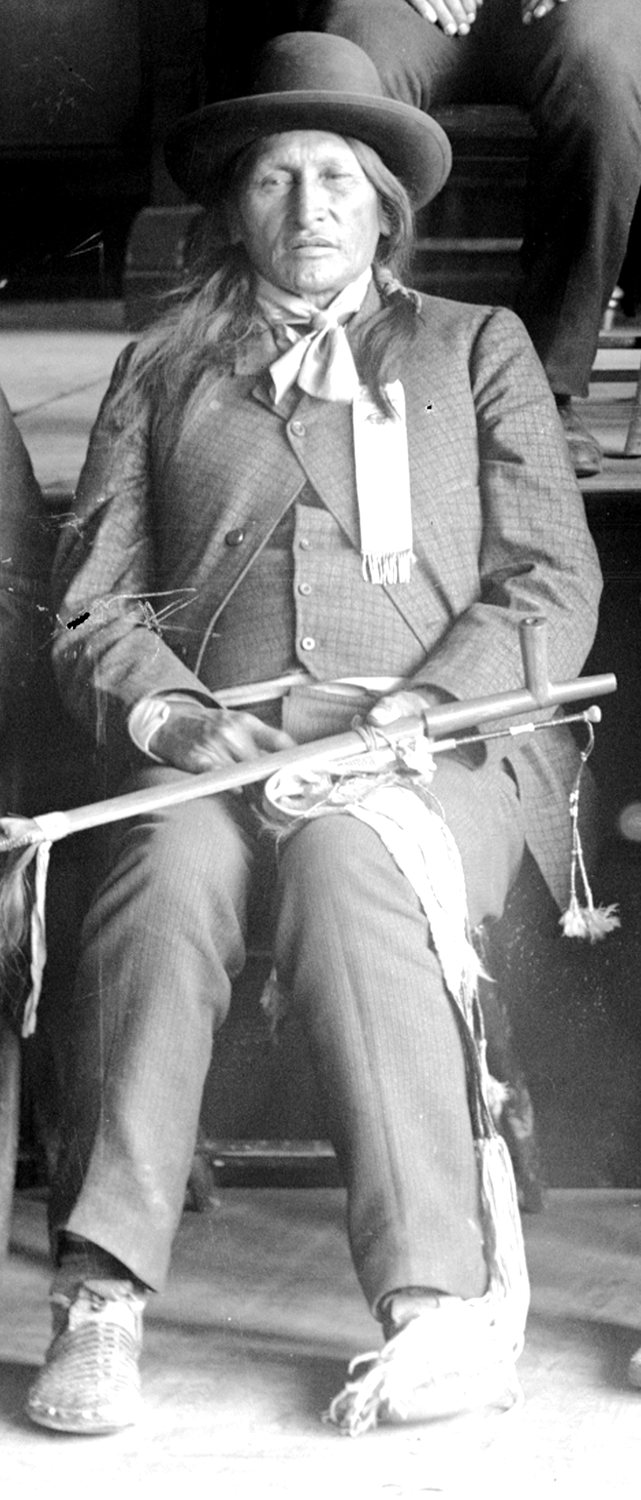 Seated portrait of Spotted Elk in a dark suit and broad-brimmed hat, holding a pipe