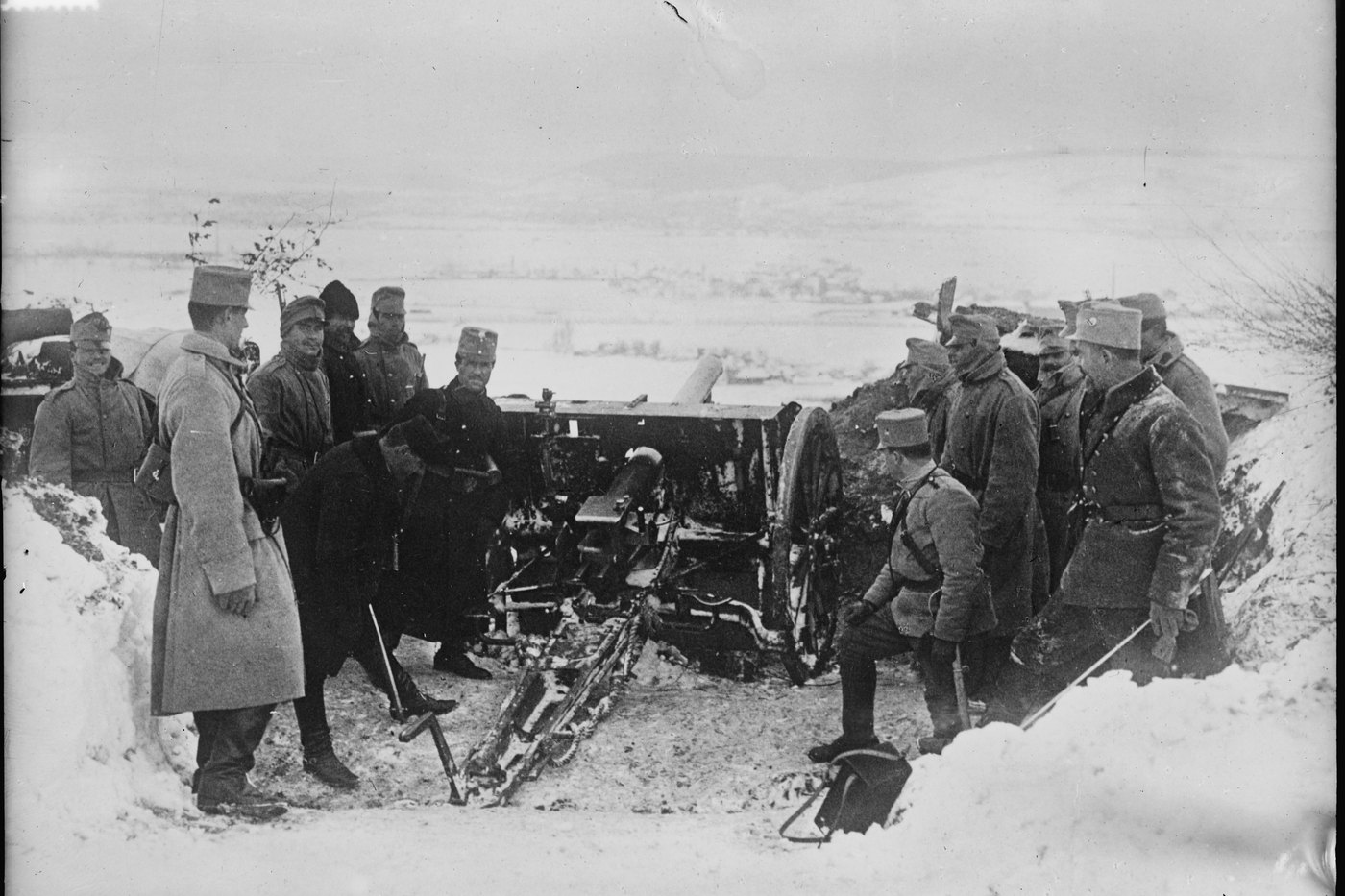 Black and white photograph: officers and gunners in greatcoats and soft caps stand around a field gun in a snow-banked position above a wide, snowy valley.