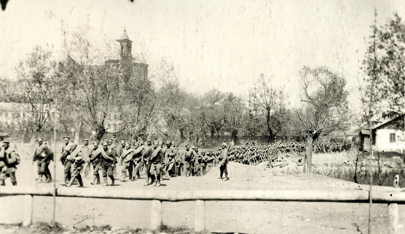 Black and white photograph: a long column of soldiers with bedrolls slung across their chests marches along a dusty road past trees and a church tower, watched by a single guard.
