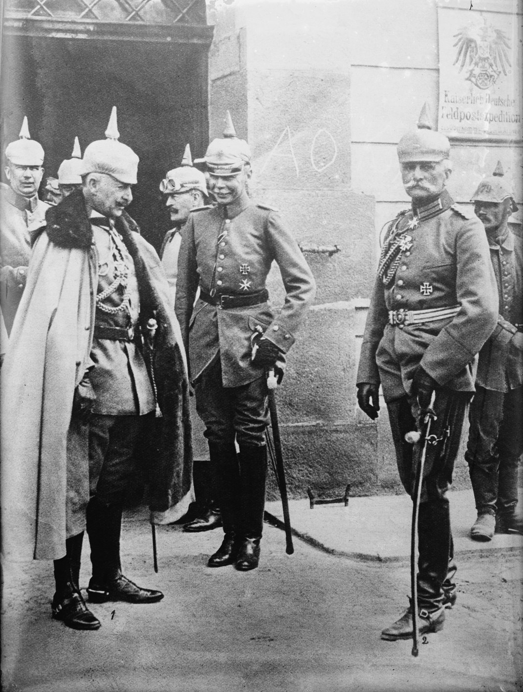 Black and white photograph: Kaiser Wilhelm II in a fur-collared cape and spiked helmet stands in a doorway facing a tall general with a white moustache, a sabre and a chest of medals, with officers behind them.