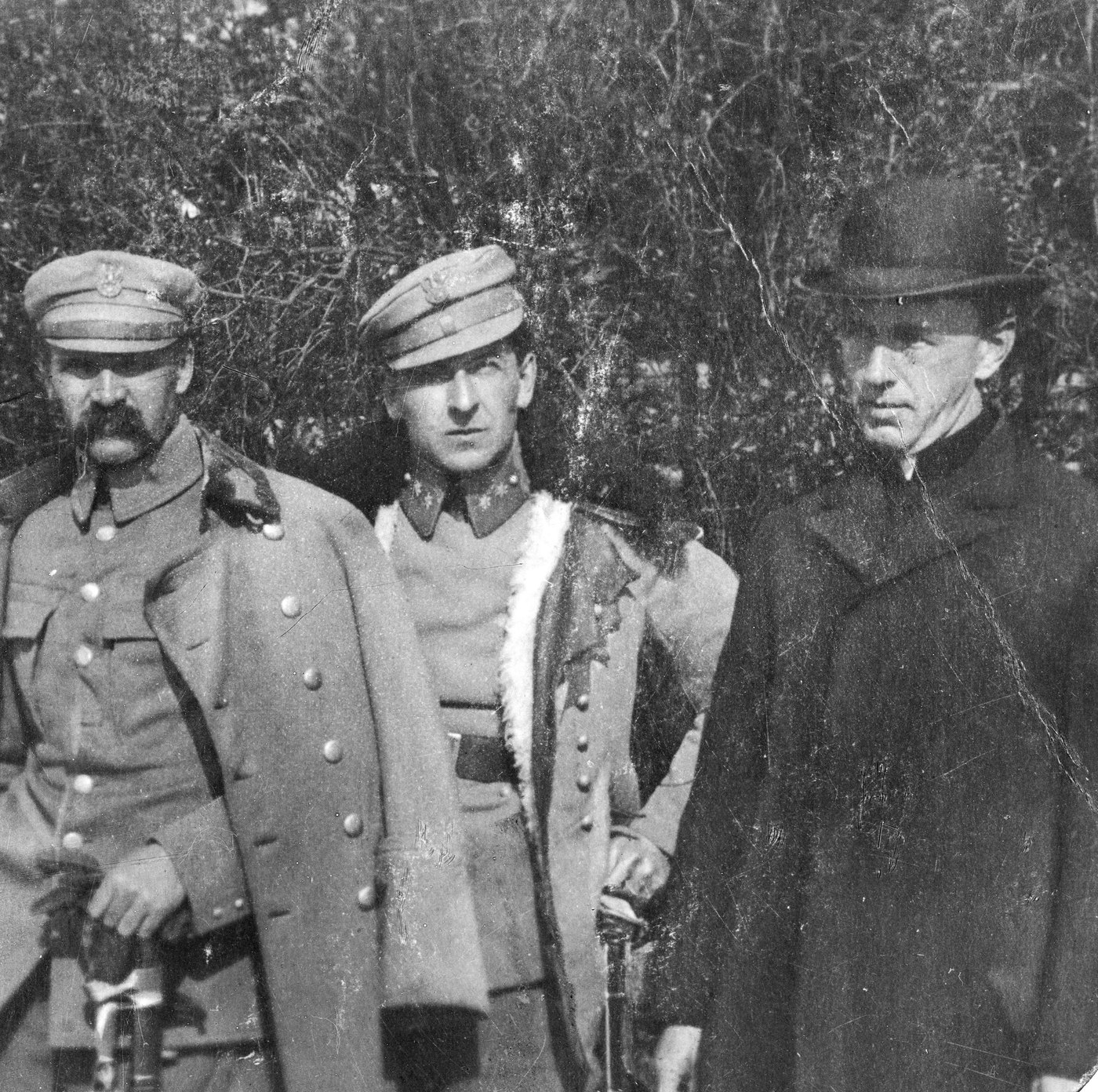 Black and white photograph: a moustached officer in a peaked cap and open greatcoat, a younger officer in a fur-lined coat and a priest in a bowler hat stand before a thicket.