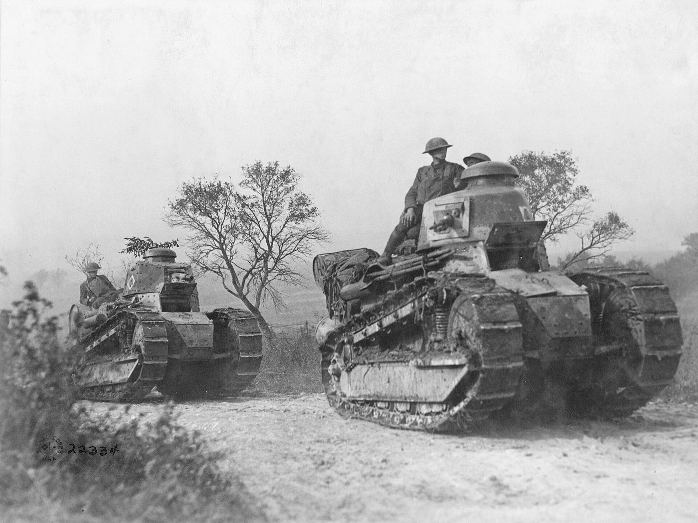 Black and white photograph: two small French-built tanks with round turrets drive along a dusty road past bare trees, American soldiers in steel helmets riding on top.