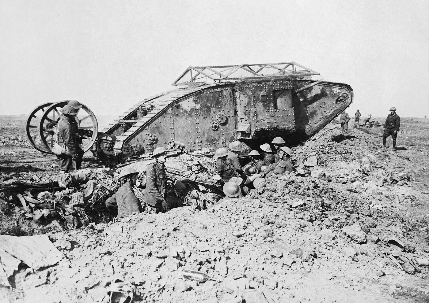 Black and white photograph: a large rhomboid British tank with a wire frame on its roof and a pair of wheels at its tail stands beside a trench full of resting soldiers in steel helmets.