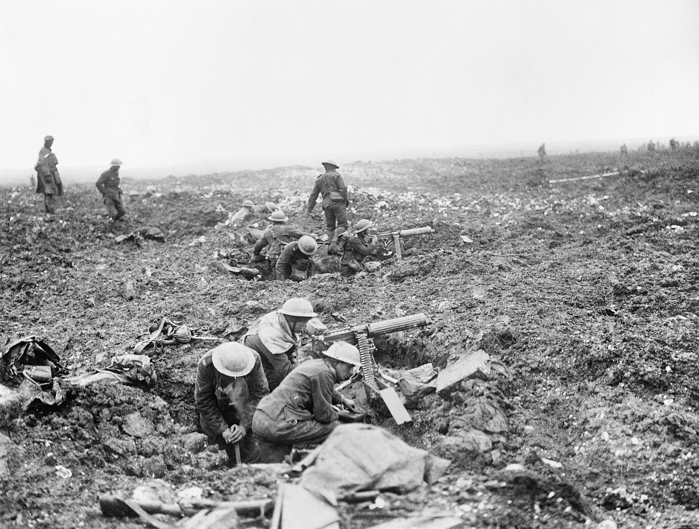 Black and white photograph: Canadian soldiers in steel helmets crouch with heavy machine guns in shell holes on a churned, open plateau, while others walk across it in the background.