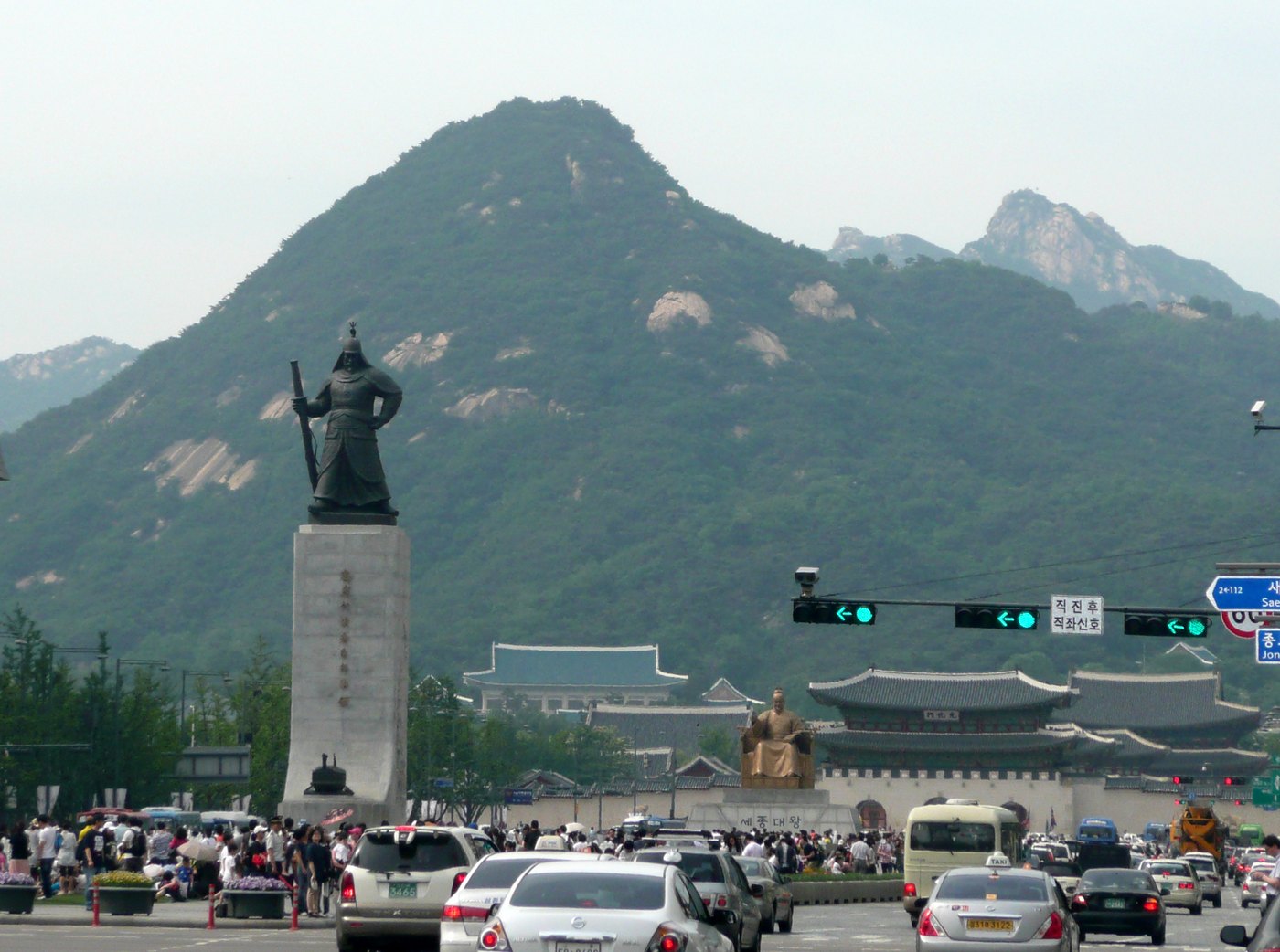 Bronze statue of an armored Korean admiral holding a sword on a tall stone plinth in a busy Seoul boulevard, with a palace gate and a green mountain behind