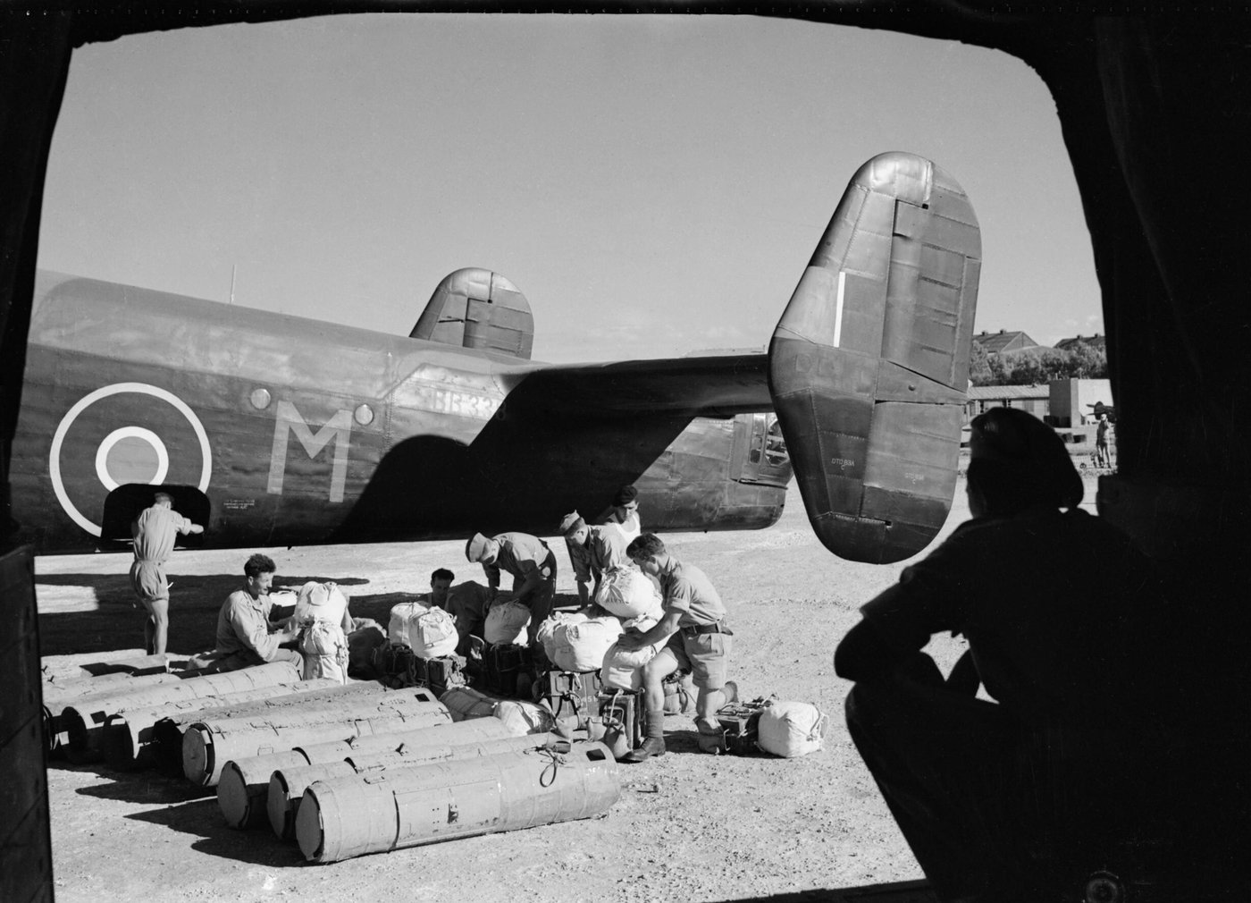Black and white photograph framed by an aircraft doorway: men loading cylindrical parachute containers and bundles beside the tail of a four-engine bomber marked M.